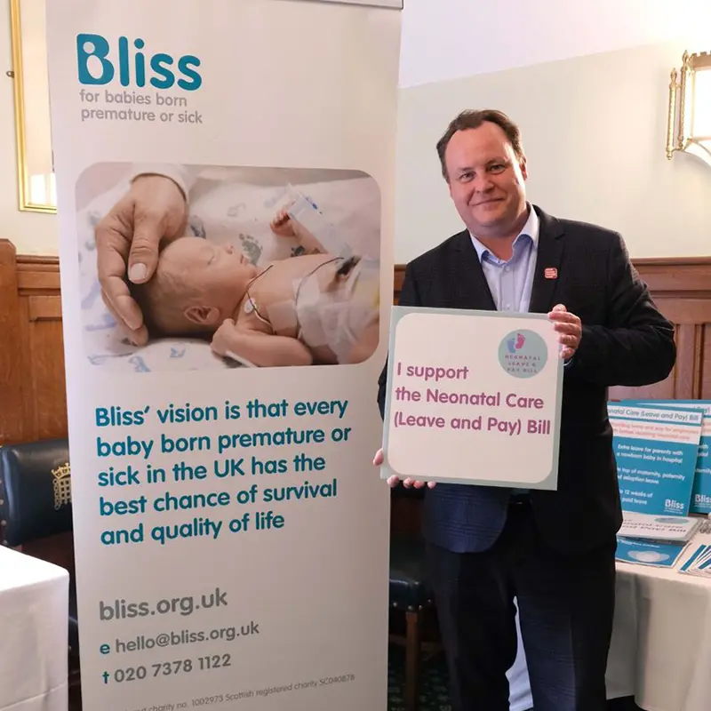 Chris Stephens MP standing holding a sign saying 'I support the Neonatal Care (Leave and Pay) Bill. Behind him is a pop up stand with a picture of a small baby in Neonatal Care, someone is stroking the babies head. Above the picture is a logo 'Bliss for babies born premature or sick in the UK has the best chance of survival and quality of life.' The contact details are bliss.org.uk email hello@bliss.org.uk tel 020 7378 1122